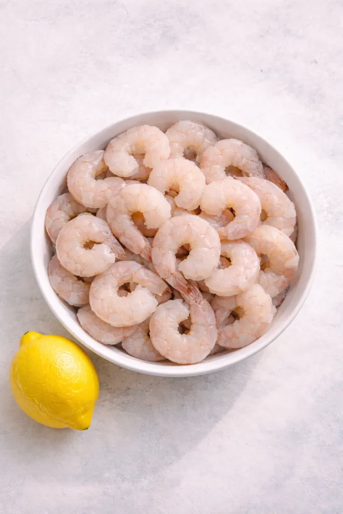 Raw peeled shrimp arranged in a white ceramic bowl with a whole lemon placed beside it on a light textured surface.